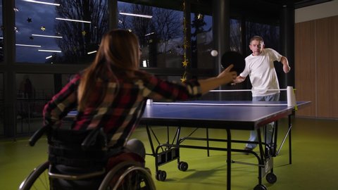Mature man with cerebral palsy and woman in wheelchair enjoying evening indoor activity while playing ping-pong game in sport hall/ Healthy, positive, active, lifestyle for persons with disabilities
