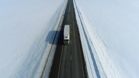 Semi trucks speed traffic driving / traveling on straight highway through snow field at winter frozen day / aerial top down view
