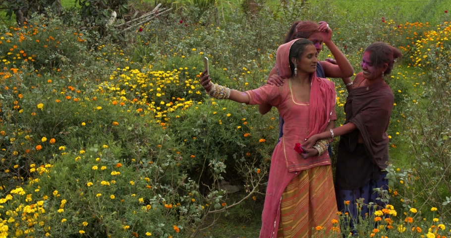 Funny Indian ladies, traditional costume jewelry, smiling joyful laugh have fun taking photos selfie video and checking it on smart phone mobile device outdoor flower garden celebrating Holi festival