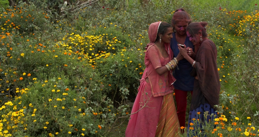 Funny Indian ladies, traditional costume jewelry, smiling joyful laugh have fun taking photos selfie video and checking it on smart phone mobile device outdoor flower garden celebrating Holi festival