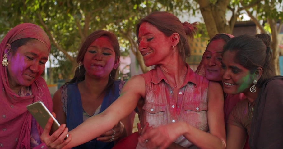 Indian women, traditional costume jewelry, smiling joyful laugh have fun taking photos selfie video with white western caucasian woman and check it on smart phone mobile outdoor in garden sitting Holi