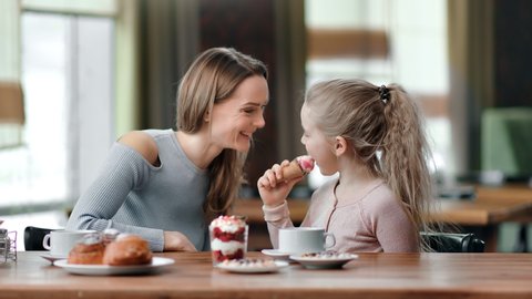 Happy mother and daughter eat dessert and drink tea together at cafe having fun. Playful kid girl licking ice cream smearing to mom nose laughing have positive emotion. Medium shot on RED camera