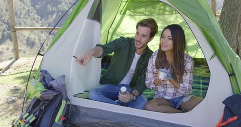 Young happy tourists sitting in tent and drinking tea while having rest in mountains.