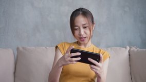 Portrait of Attractive Asian Woman using smartphone playing a video game on mobile having fun and happy emotion sitting on couch in the living room at home. Relax and Leisure time concept.