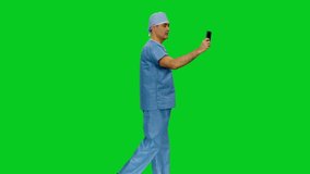 Asian mid adult doctor in uniform having a video chat while walking against green screen background, Side view, Full HD chroma key footage