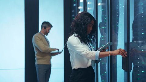 Afro-american young IT specialist woman using digital pad opening rack door of server cabinet analyzing statistics information cooperating in data center.