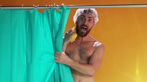 Young bearded man wearing cap washing himself in the shower looking scared of being peeped. Male silhouette reflecting on shower bath curtain. Comical scene.
