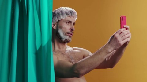 Sexy young attractive man with soap beard making funny face grimaces on camera taking selfies smartphone taking shower at home. Comedy. Comical scene.