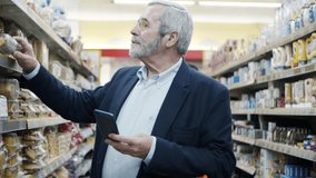 Mature man using smartphone in supermarket. Focused bearded man holding mobile phone and choosing goods in grocery store, handheld shot. Shopping and technology concept
