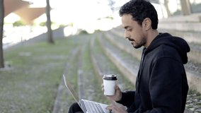 Man using laptop and drinking coffee to go. Side view of focused young man using laptop computer and drinking coffee from paper cup on street, handheld shot. Freelance concept