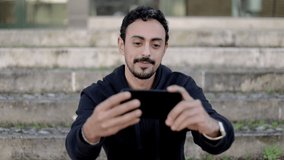 Cheerful man having video chat on street. Handsome happy young man sitting on steps, waving hand and talking during video call outdoors. Communication concept