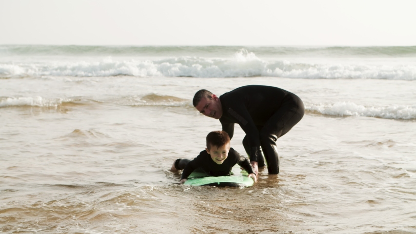 Adorable happy boy swimming on board. Happy father helping little son in wetsuit lying and swimming on surfboard on waves, handheld shot. Surfing concept