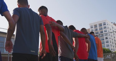 Low angle rear view of a multi-ethnic group of male football players in casual sportswear, standing in a row on a playing field on a sunny day, embracing, holding a football, in slow motion
