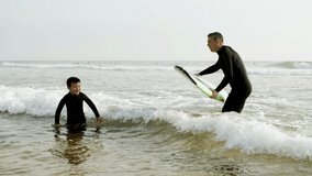 Father teaching son surfing in ocean. Happy father helping little son in wetsuit lying and swimming on surfboard on waves. Surfing concept