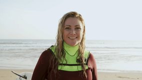 Happy female surfer smiling at camera. Front view of cheerful middle aged woman in wetsuit holding surfboard and looking at camera on sea coast, handheld shot. Water sport concept