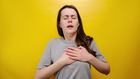Unhealthy woman feeling pain ache touching chest having heart attack, worried girl suffers from heartache, infarction or female heart disease and arrhythmia concept, isolated on yellow background