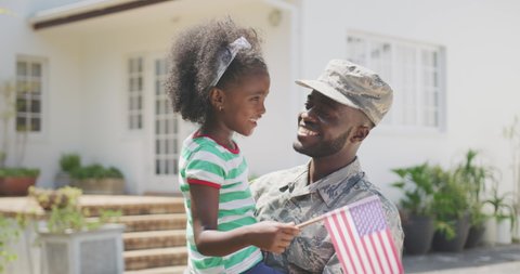 Front view of a happy African American man enjoying time in the garden, with his family, wearing military uniform, holding up his African American daughter holding a flag, on a sunny day