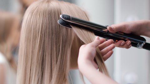 Caring hands of professional female hairdresser straighten blonde hair using hot iron tool. Artist woman arms making straightening hairdo at beauty salon. Close up shot on 4k RED camera