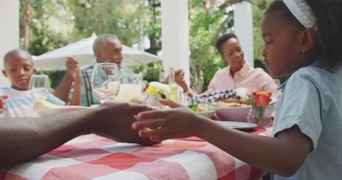 Side view of a multi-generation multi-ethnic family having a good time in a garden, sitting by a table with food on it, holding hands, saying grace on a sunny day, in slow motion