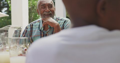 Over the shoulder view of a senior mixed race man having a good time with his family, sitting by a table in a garden, talking to his grandson, pointing at him, smiling, in slow motion