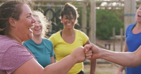 Side view close up of two happy Caucasian women laughing and fist bumping, with a multi-ethnic group of female friends in the background enjoying exercising at boot camp together, relaxing after
