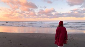 Carefree woman dressed in red shirt and hat walking on the beach at dusk. View from the backside. Life enjoyment on nature concept. Video recorded on a smartphone