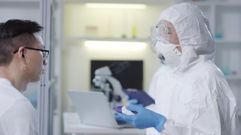 Tracking of medical scientist in disposable coveralls, mask and goggles using forehead thermometer and checking temperature of contagious male clinical trial participant, then talking to colleague