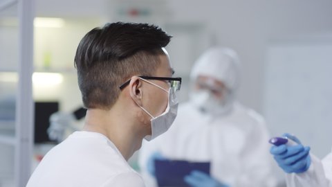 Tracking of doctor in disposable coveralls and gloves using forehead thermometer and checking temperature of contagious patient in mask. Medical scientist conducting research in background