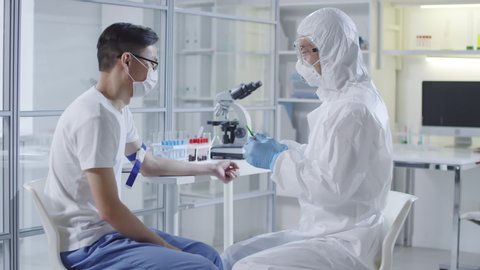 Tracking of unrecognizable doctor or medical scientist in protective suit, gloves, mask and goggles injecting sick Asian patient with experimental vaccine from coronavirus