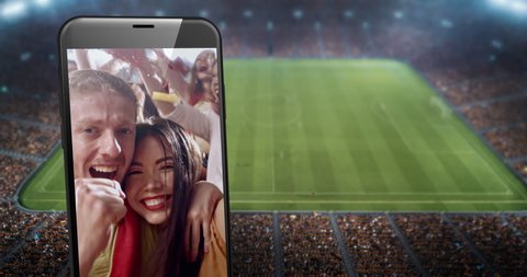 A phone with video of cheering fans on the screen is placed in front of professional soccer stadium. Stadium is made in 3D.