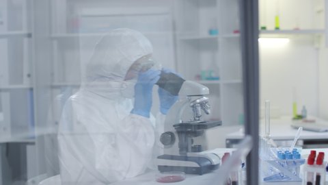 Tracking of female medical scientist in hazmat suit, respirator mask, gloves and goggles looking into microscope, then inspecting tube with blood sample while conducting research in laboratory