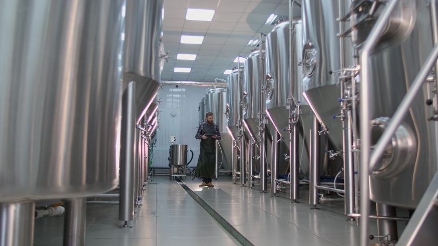 A male brewer with a beard goes to the beer-producing room and will check the data of the devices using a tablet