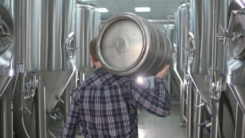 A working male brewer carries a keg filled with beer as he passes beer tanks. close-up. back view