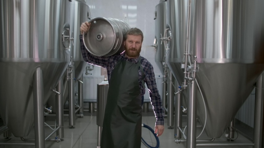 Worker Male brewer carries a keg filled with beer passing beer tanks | Shutterstock HD Video #1047821842