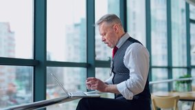 Thoughtful middle aged businessman in suit with a laptop sitting near the window working with documents, typing. Business video.