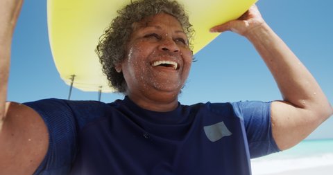 Low angle front view close up of a senior African American woman on a beach in the sun, holding a surfboard on her head, with blue sky and calm sea in the background in slow motion