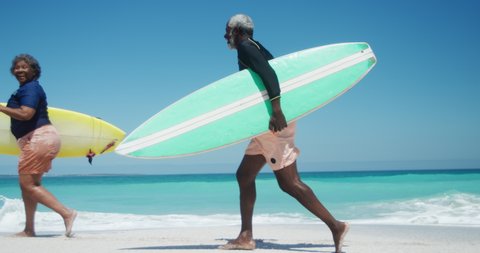 Side view of a senior African American couple on a beach in the sun, holding surfboards under their arms and running, with blue sky and sea in the background in slow motion
