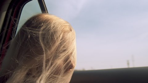 Woman looks out open car window as blond hair swirls across face, close-up