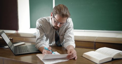 Bearded teacher in eyeglasses checking tests in classroom, modern education