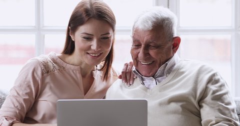 Smiling young adult grown granddaughter teaching old elder grandfather using laptop computer explaining device applications work sit on sofa at home, senior person learning modern technology concept