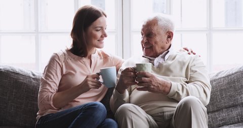 Happy senior elder father grandfather and young adult granddaughter grown daughter talking bonding drink tea sit on sofa, loving 2 two generations family embracing enjoy friendly conversation at home