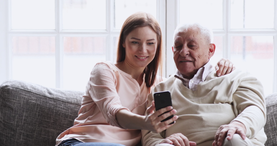 Cheerful young adult grownup granddaughter laughing watching funny social media videos using smart phone sit on sofa embracing teaching elder grandpa older generation having fun relaxing with gadget Royalty-Free Stock Footage #1047837424