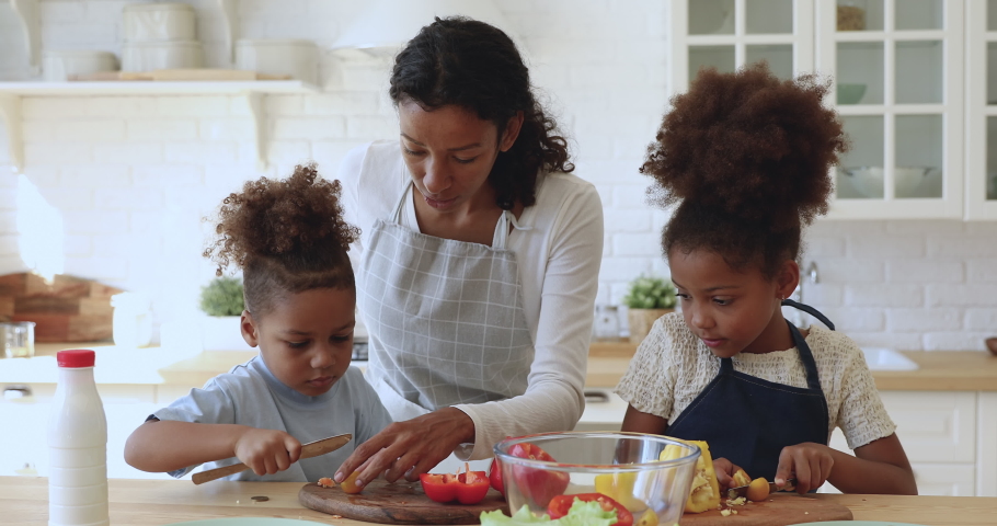 Happy attractive young african american mom in apron teaching little kids siblings cutting fresh vegetables for vegetarian food. Smiling family of three preparing food together at modern kitchen. Royalty-Free Stock Footage #1047837448