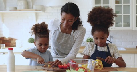 Happy attractive young african american mom in apron teaching little kids siblings cutting fresh vegetables for vegetarian food. Smiling family of three preparing food together at modern kitchen.