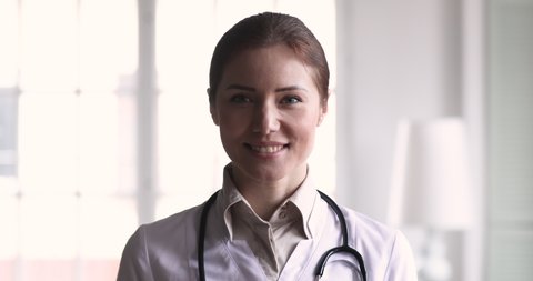 Smiling confident young adult female doctor close up portrait, friendly happy woman physician or nurse professional general practitioner posing with stethoscope looking at camera in medical office