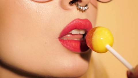Closeup red lips of young beautiful hipster bad girl with earring in her nose.Sexy carefree smiling woman posing in studio near beige wall. Positive model licking round candy lollipop
