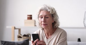 Smiling senior adult grandmother using smartphone sitting on sofa. Happy 70s elder woman holds mobile phone texting message, checking app, reading news at home. Old grandparent learns tech gadget