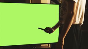 Man with Remote Control over a Green Screen TV. Fast Zoom In. You can Replace Green Screen with the Footage or Picture you Want with “Keying” effect in After Effects (check out tutorials on YouTube).