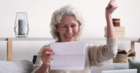 Cheerful old senior woman reads postal mail approval letter excited by good news. Excited elder lady feels satisfied with bill or tax refund, bank service offer, holding paper sitting on sofa at home