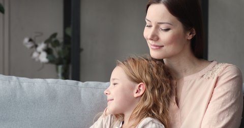 Loving single mother embracing kid daughter looking away. Young smiling foster care parent mum hugging adopted child girl sitting on sofa dreaming of good future enjoying sweet moment of love concept.